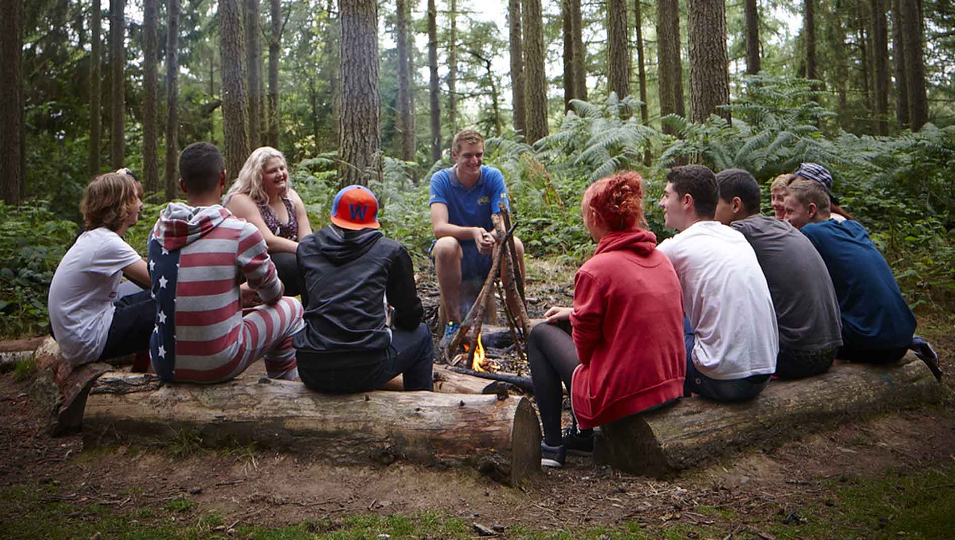 A group of people sit on logs around a campfire in a wooded area, engaging in conversation.