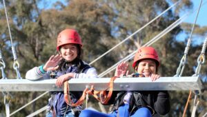 Two children in red helmets smile and wave while sitting on a high ropes course. Trees and ropes are visible in the background.