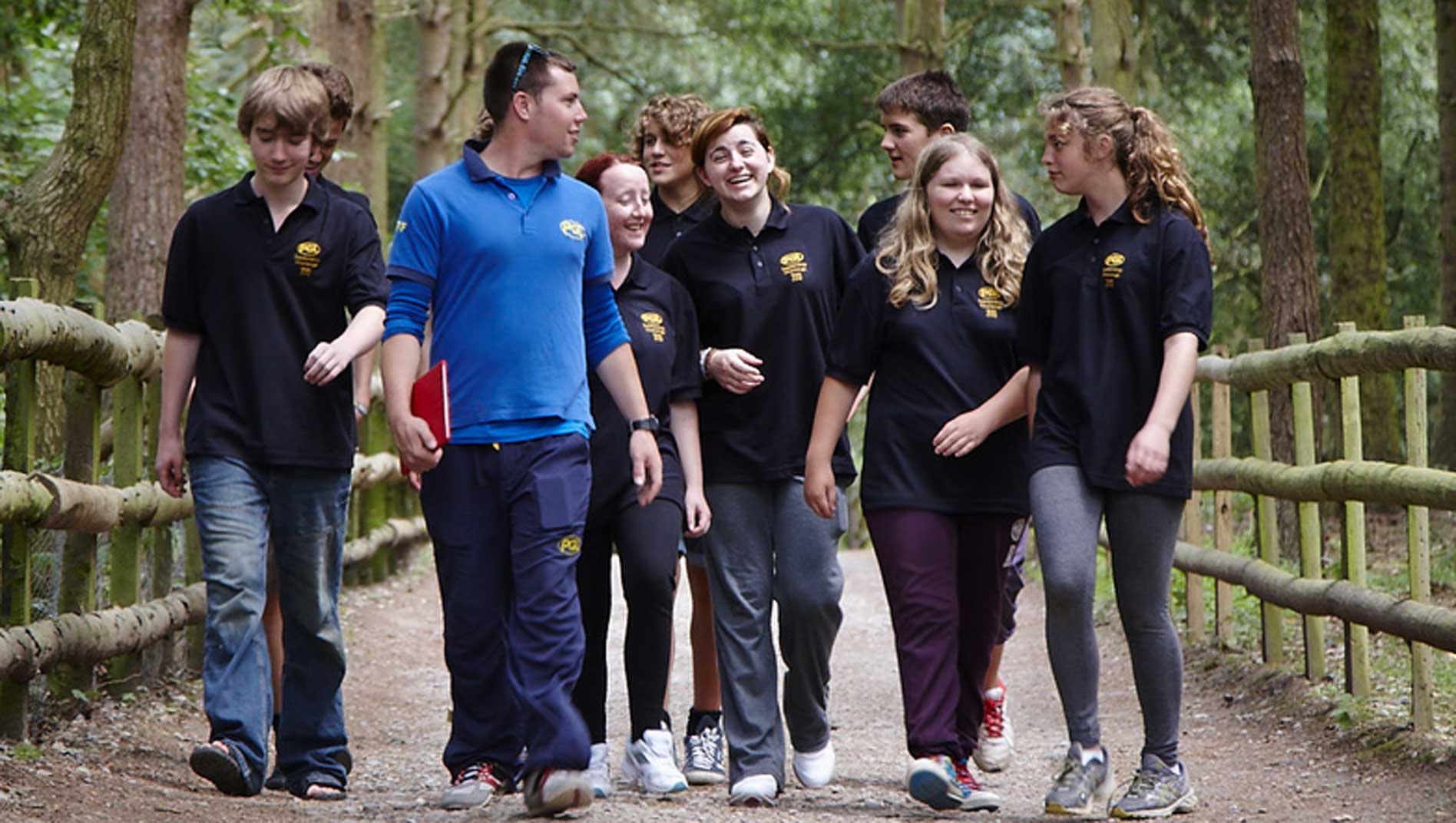 A group of people, wearing matching black shirts, walk with a leader in a blue shirt along a wooded path, surrounded by trees and a wooden fence.