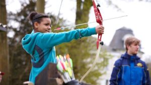 A youth wearing a blue hoodie is drawing a red bow to shoot an arrow, with a quiver of arrows visible in the foreground. Another person is standing in the background wearing a blue jacket.