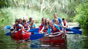 A group of children and adults wearing life jackets paddle red canoes on a calm river surrounded by green trees.