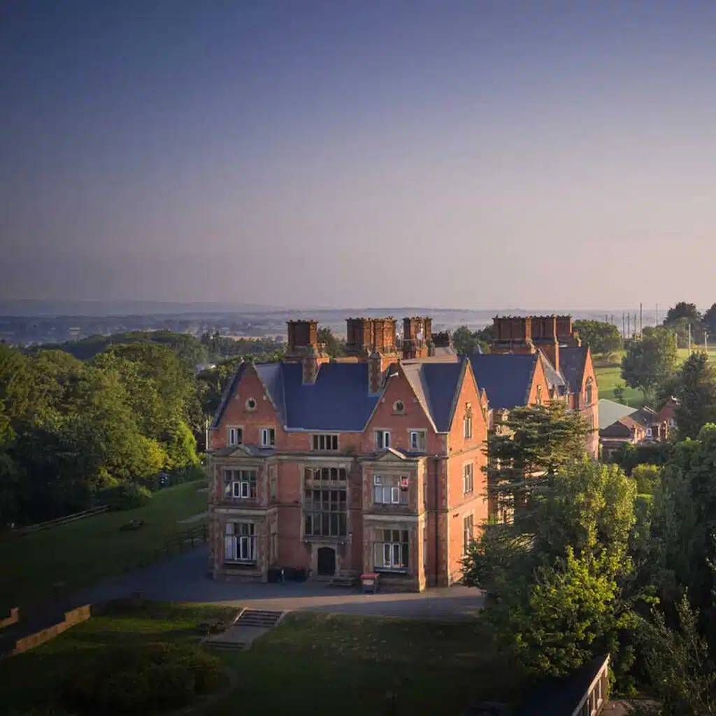 A large brick manor house with multiple chimneys, surrounded by greenery and trees under a clear blue sky, reminiscent of the idyllic settings often found at PGL camps.