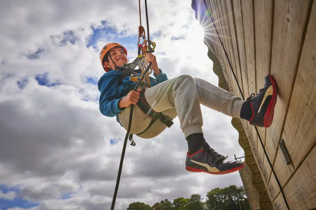 A child wearing a helmet and harness abseils down a wooden wall on a sunny day, gripping climbing ropes and smiling—an exciting moment preparing for your child’s first school trip away from home.