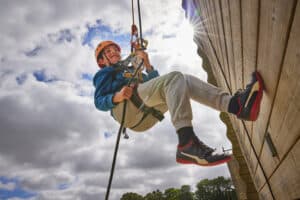 A child wearing a helmet and harness abseils down a wooden wall on a sunny day, gripping climbing ropes and smiling—an exciting moment preparing for your child’s first school trip away from home.