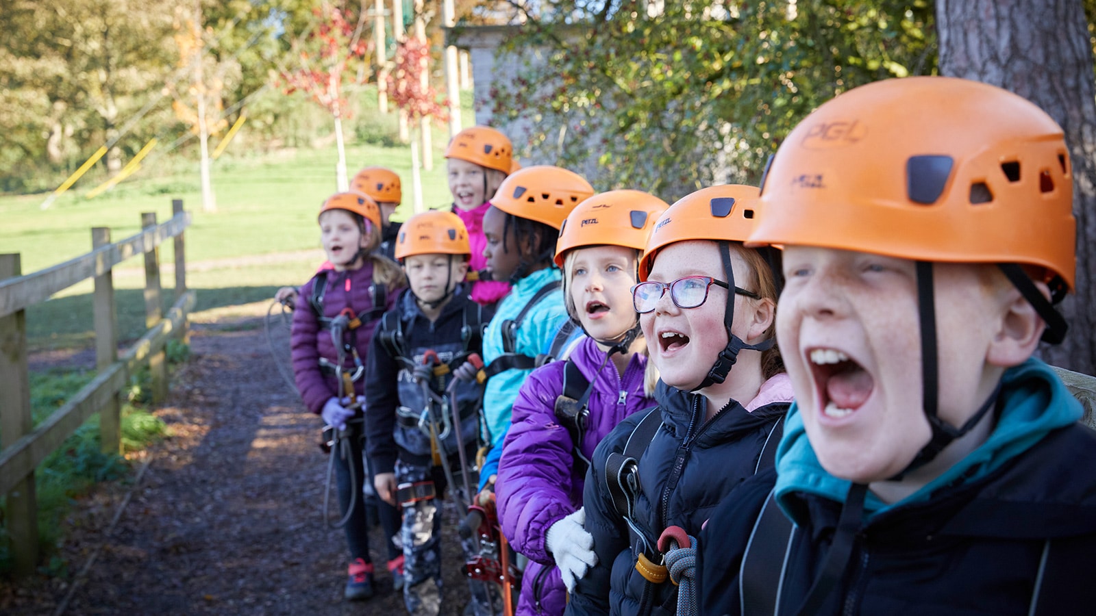 A group of children wearing helmets and outdoor gear stand in a line, appearing excited and shouting, while partaking in an outdoor adventure activity.