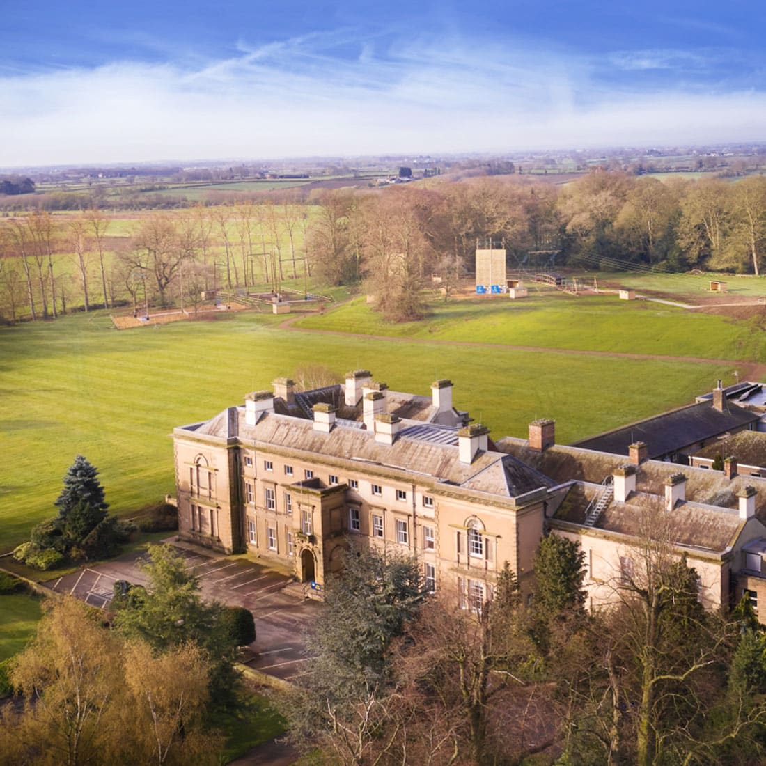 An aerial view of a large historic building, reminiscent of PGL camps, surrounded by a vast green lawn and trees under a blue sky.