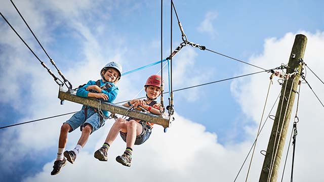 Two children wearing safety harnesses and helmets sit on a suspended swing against a blue sky with clouds, enjoying the thrilling activities offered at PGL camps. They are securely fastened by ropes attached to a wooden pole.