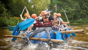 Five people in helmets and life jackets paddle homemade rafts in a lake surrounded by trees, enjoying the adventure typical of PGL camps.