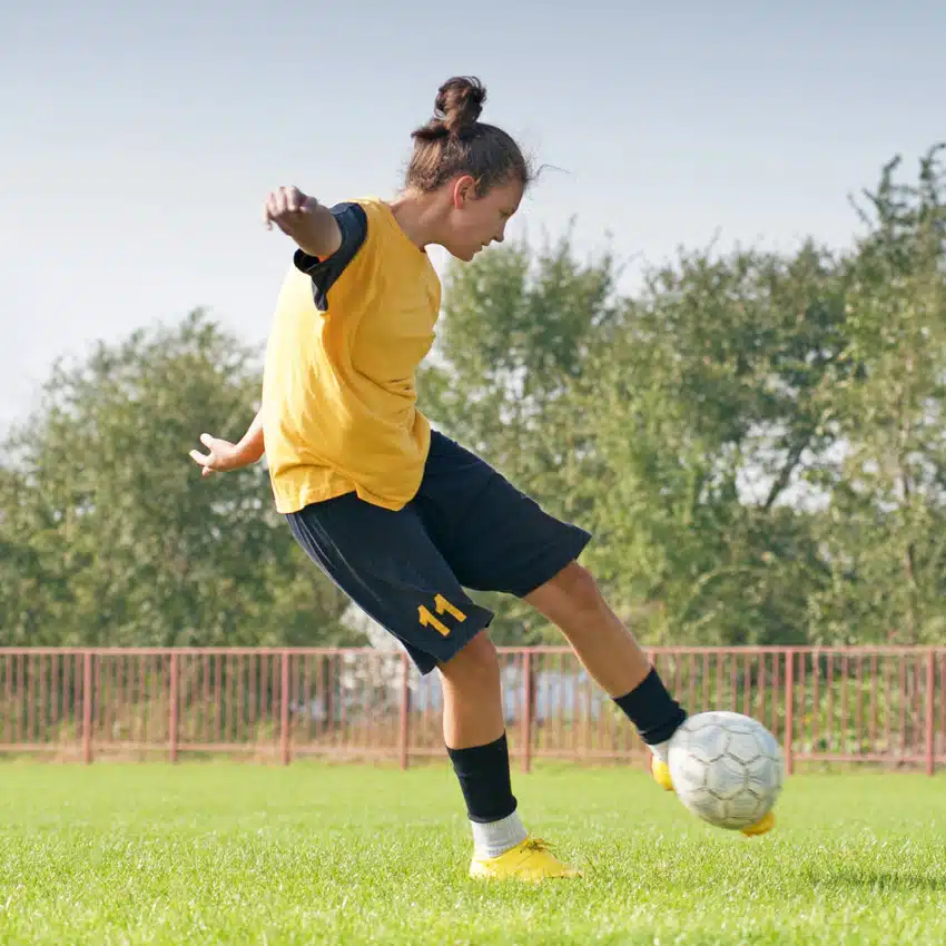 A soccer player wearing a yellow jersey and black shorts with the number 11 is kicking a soccer ball on a grassy field near Newby Wiske Hall. Trees and a fence are visible in the background.