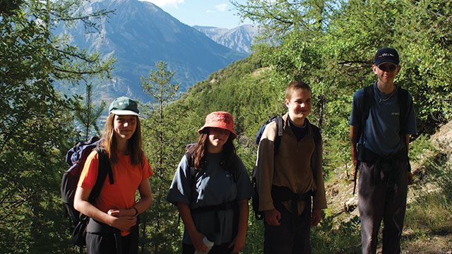 Four hikers stand on a trail with mountains and trees in the background, each dressed in casual outdoor attire and carrying backpacks.