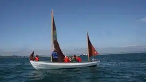 A small sailboat with a group of people wearing life jackets navigating calm waters on a clear day.