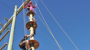 Three people in safety gear are climbing a tall wooden pole with circular platforms, using ropes for support, against a clear blue sky.