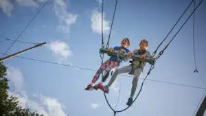 Two children wearing safety harnesses are seated on a high swing, suspended in the air against a blue sky with some clouds.