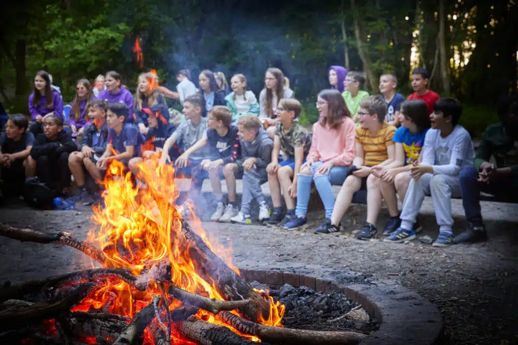 A group of people, mostly children, sit on logs in a forest around a large campfire, watching it intently.