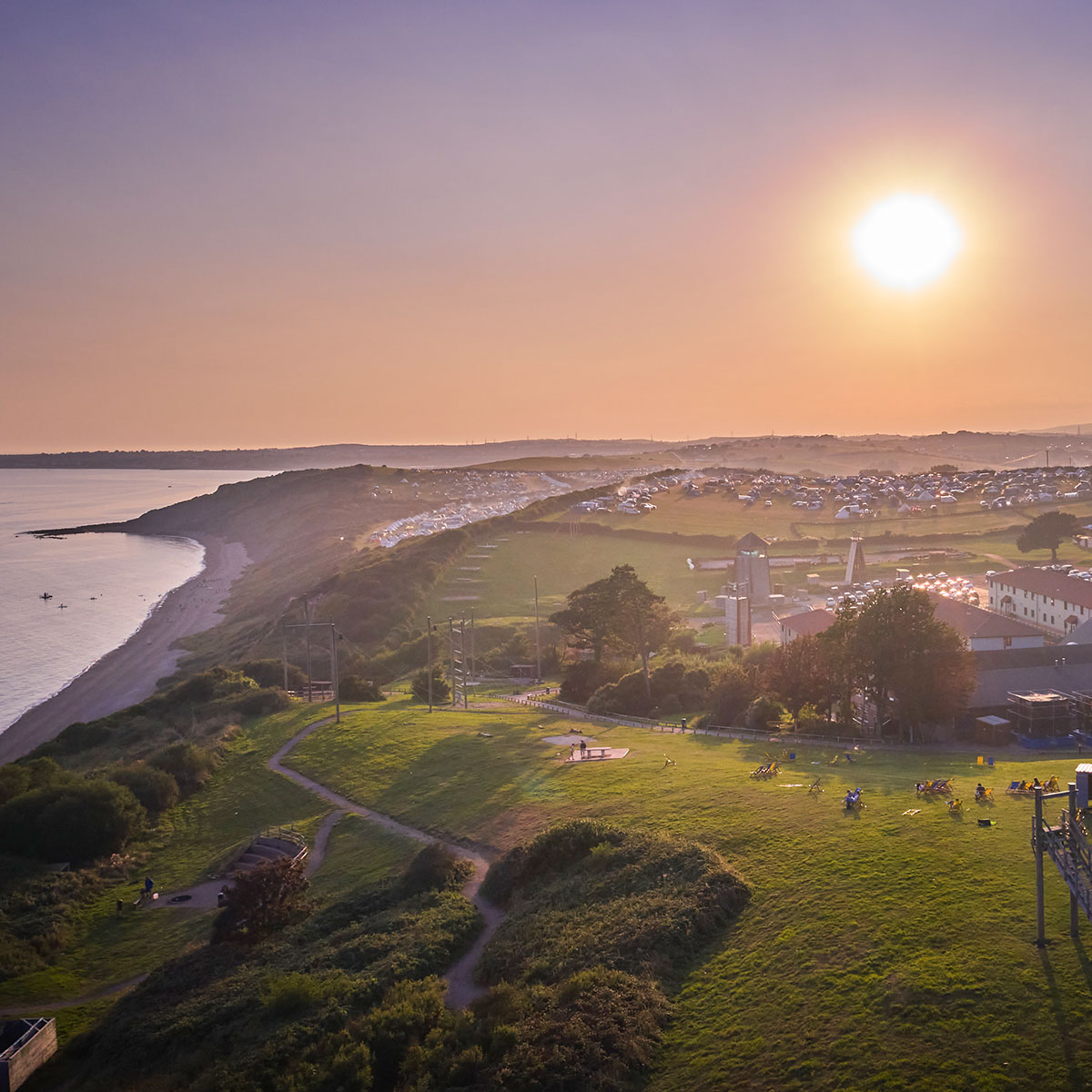 A coastal landscape at sunset with lush green fields, scattered buildings, and tents. The sun is low in the sky, casting a warm glow over the scene. Hills and the sea are visible in the background.
