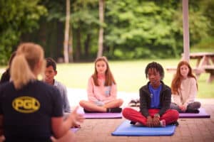 Children sitting cross-legged on yoga mats outdoors during a class, with a leader wearing a PGL shirt. Trees and a picnic table are in the background.