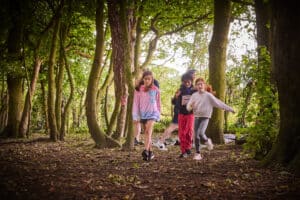 Children walking through a forest, surrounded by tall trees with green foliage.