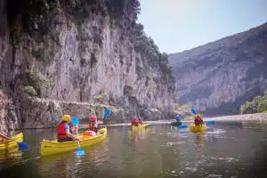 A group of people wearing life jackets paddle yellow canoes on a calm river, surrounded by steep rocky cliffs and green hills.