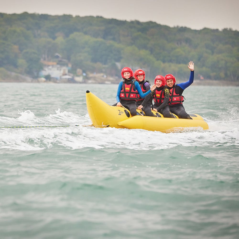 Four people wearing helmets and life vests ride a banana boat on a body of water with forested land in the background. One person waves while the others look ahead.