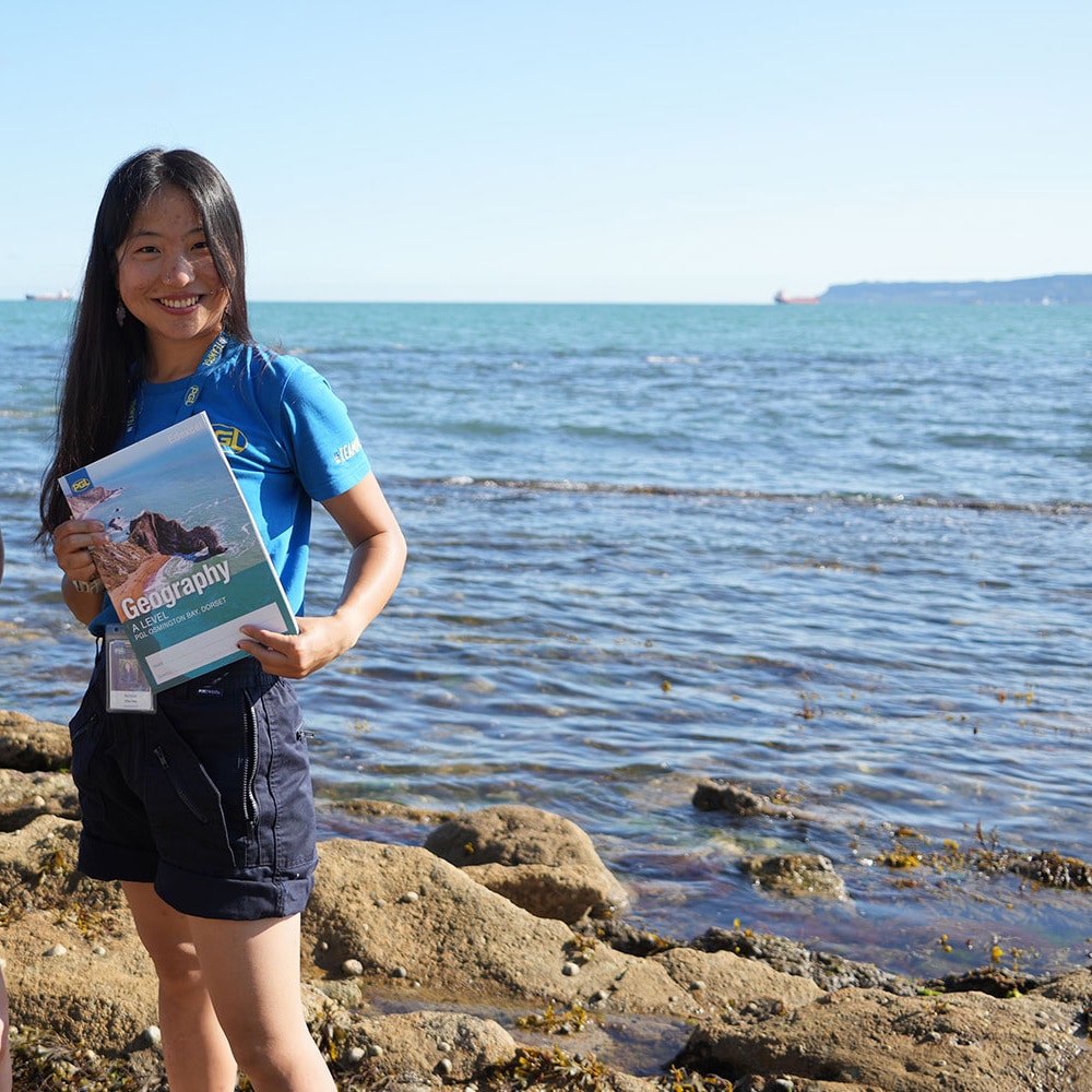 A woman stands on rocky terrain by a body of water, smiling and holding a geography book. She is wearing a blue shirt and shorts.