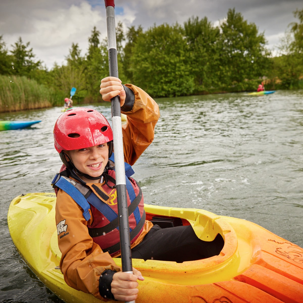 Person in a red helmet and orange jacket kayaking on a calm body of water with trees in the background.