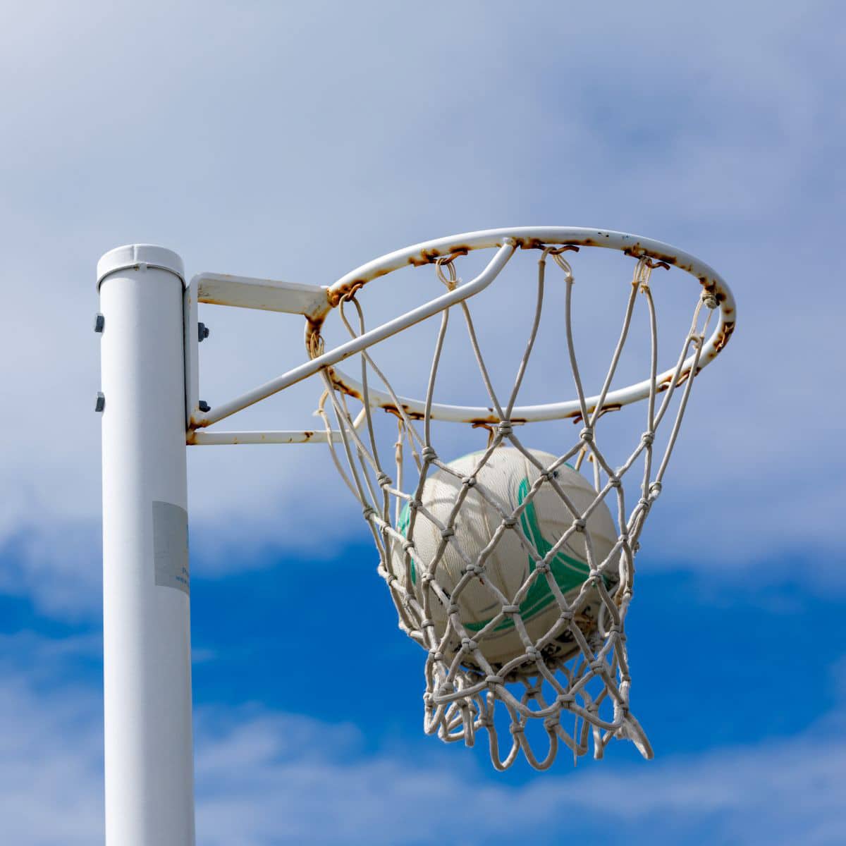 A white and green ball stuck in a rusted netball hoop against a blue sky with some clouds.