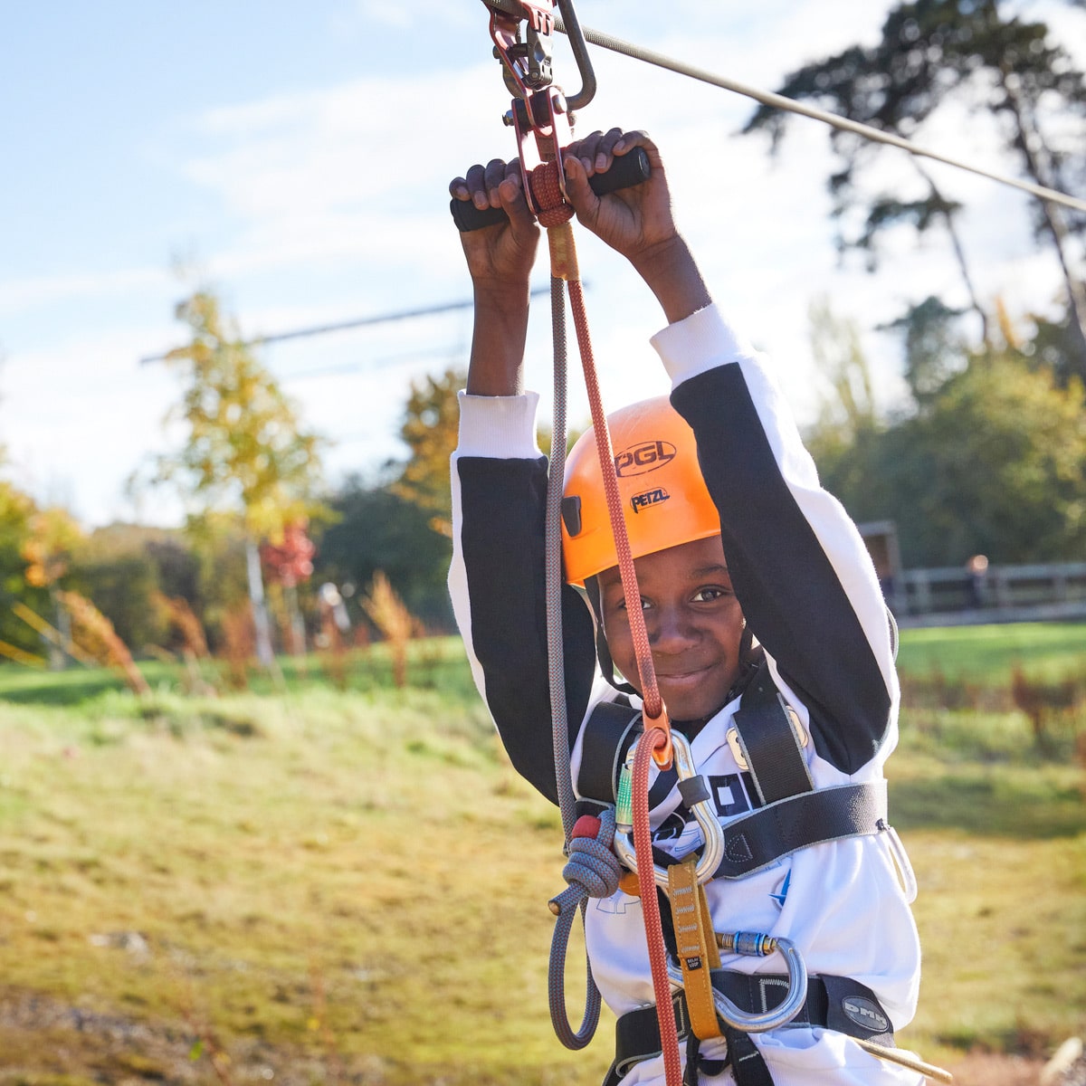 A person wearing a helmet and safety harness is holding onto a zip line while smiling. The background shows an outdoor setting with trees and grass.