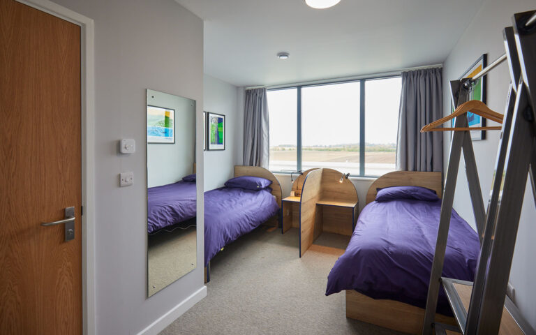 A small dormitory room with two single beds covered in purple duvets, a large window, and framed artwork on the walls. A wooden divider separates the beds, and a standing coat rack is near the corner.