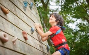 A child in a red T-shirt and safety harness climbs an outdoor wooden rock climbing wall surrounded by trees, enjoying the multi-activity adventure.