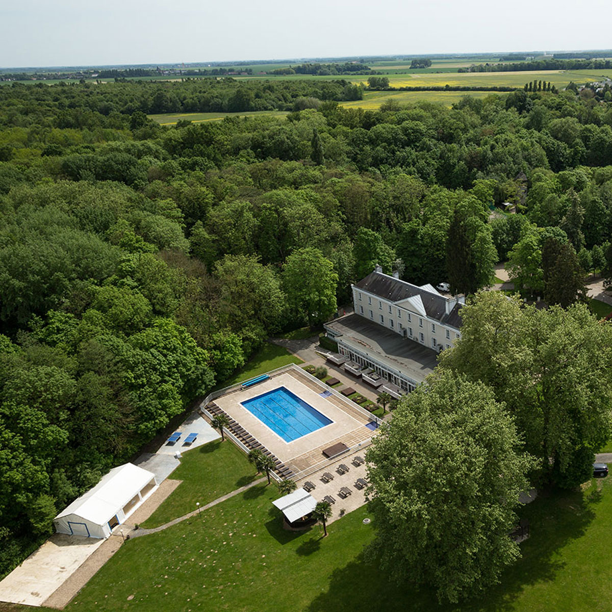 Aerial view of an outdoor swimming pool surrounded by lounge chairs, adjacent to a large house in a forested area with open fields in the background.