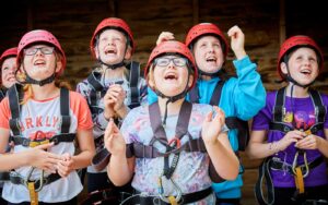A group of children wearing safety harnesses and red helmets smile and cheer while looking upwards.