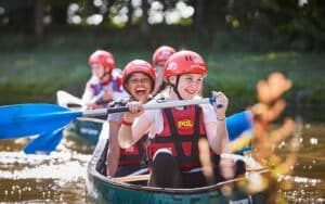 A group of young people paddles a canoe on a sunny day, wearing red helmets and life vests, smiling and enjoying the outdoor activity.