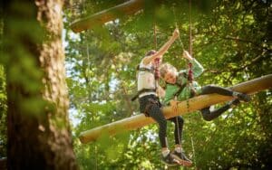 Two children wearing safety harnesses navigate a suspended wooden obstacle in a forested outdoor adventure park.