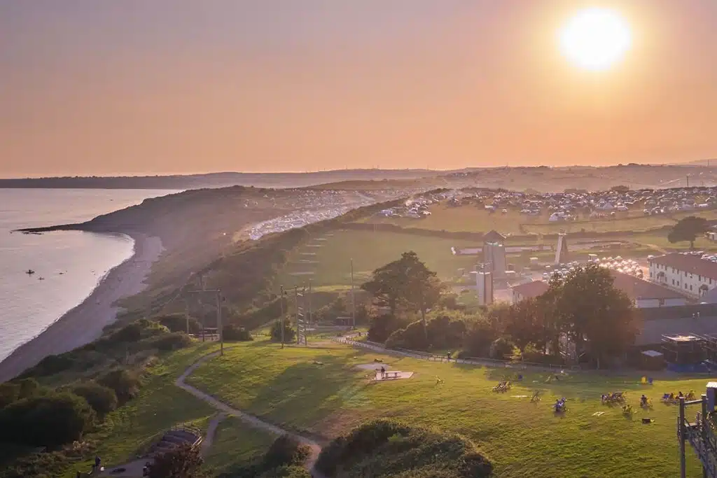 Aerial view of a coastal landscape at sunset, with a beach on the left, rolling hills, scattered buildings, and small groups of people on green fields in the foreground.