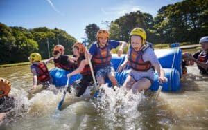 Group of people wearing life vests and helmets paddle and splash in water while on makeshift rafts during an outdoor activity on a sunny day.