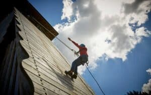 Person climbing a wooden wall on a sunny day, secured with a harness and rope, against a backdrop of clouds and blue sky.