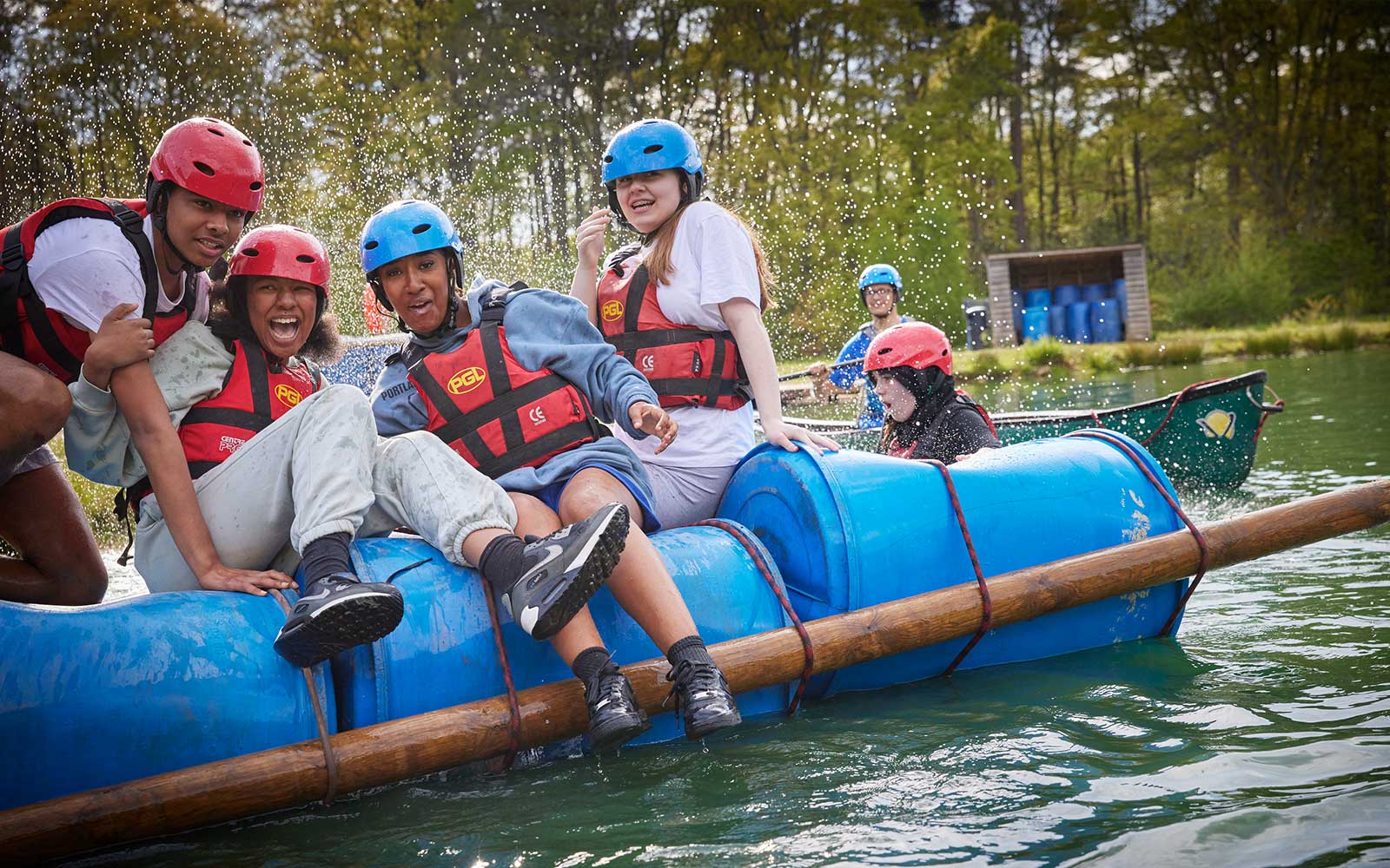 A group of people wearing helmets and life vests are on a makeshift raft in a lake, leaning and smiling as water splashes. Another raft and people are visible in the background. Forested area around the lake.