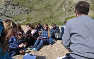 A group of people sit on a rocky hillside, some with clipboards, as a person in a grey shirt speaks to them. The setting appears to be outdoors on a sunny day, suggesting a multi-activity event.