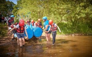 A group of people in helmets, life vests, and paddles work together to move a makeshift raft with blue barrels into the water near a grassy shore, part of an adventurous multi-activity day.