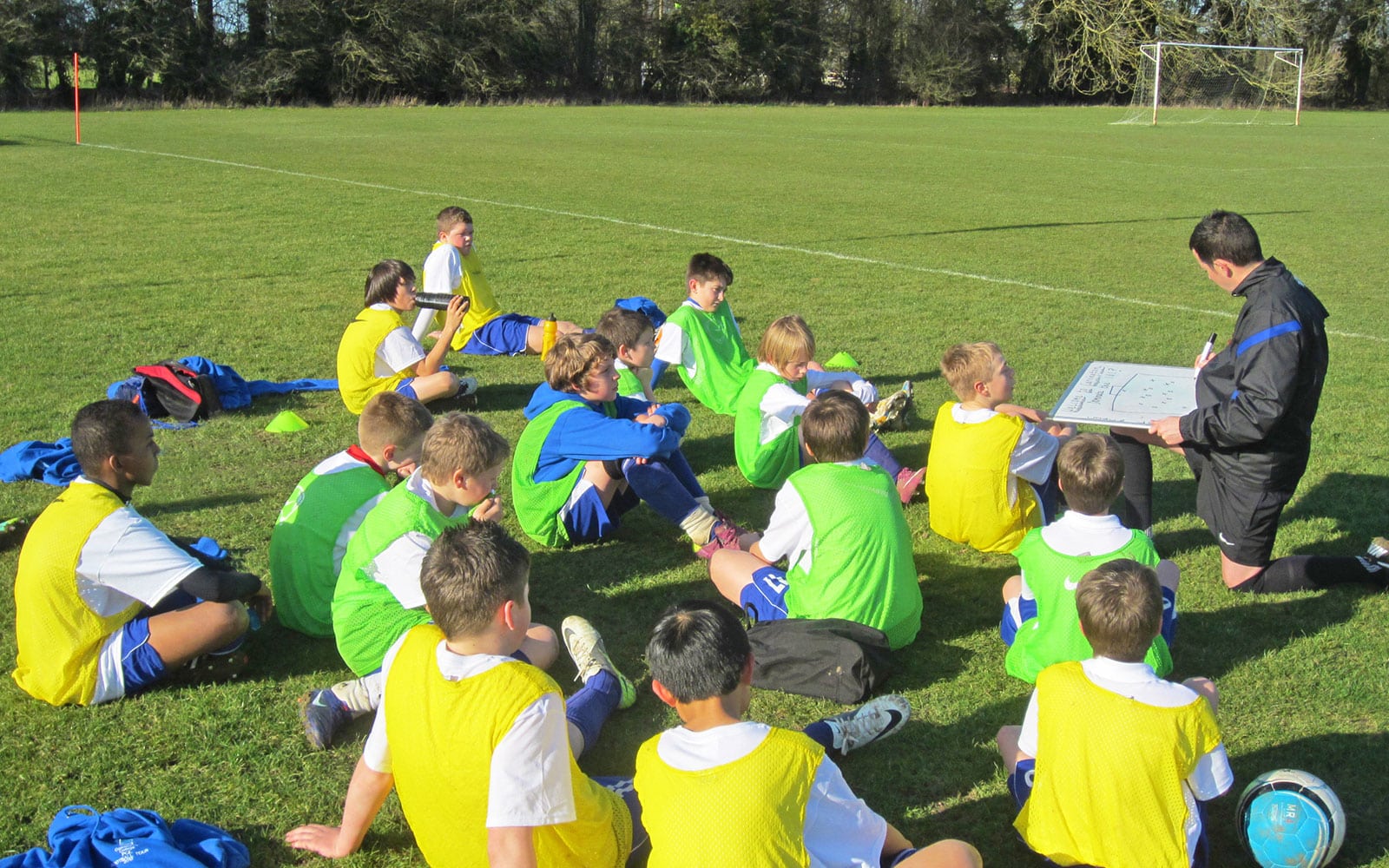 A group of children wearing soccer uniforms and yellow vests sit on the grass while a coach kneels and explains tactics using a clipboard. The multi-activity session, filled with learning and fun, ensures they're always engaged on the soccer field.