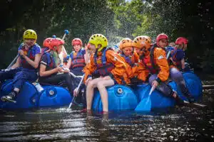 A group of children wearing helmets and life jackets sit on a raft made of blue barrels, paddling and splashing on a body of water on a bright day.