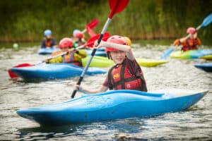 Children paddle blue kayaks on a calm lake, wearing helmets and life jackets.