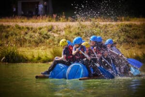 A group of people, wearing helmets and life vests, paddle a makeshift raft made of barrels on a lake, causing splashes.
