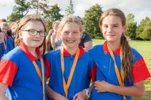 Three girls in blue and red uniforms with yellow lanyards smile at the camera while standing outside on a sunny day. Other people and trees are visible in the background.