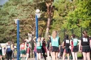 A group of girls playing netball outdoors, with one girl jumping to shoot the ball into the net. They are wearing team bibs in green, red, and blue with positional letters. Trees are in the background.