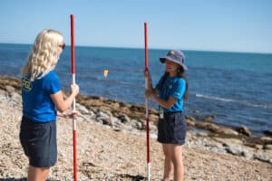 Two people in blue shirts hold measuring poles and string on a rocky beach with the ocean in the background.