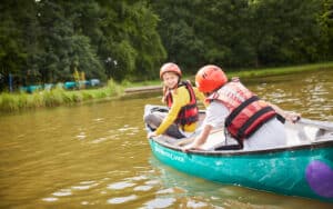 Two people in life jackets and helmets paddle a canoe on a calm lake surrounded by trees. One smiles at the camera while the other faces away.