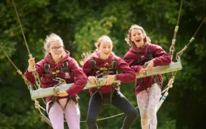 Three girls wearing harnesses and maroon sweatshirts sit together on a swing, smiling and laughing. Green trees are visible in the background.