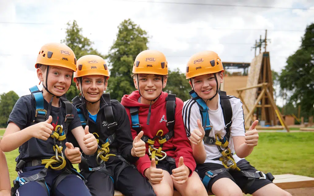 Four children wearing helmets and harnesses give thumbs-up while sitting on a bench, smiling at the camera, with a wooden climbing structure in the background.