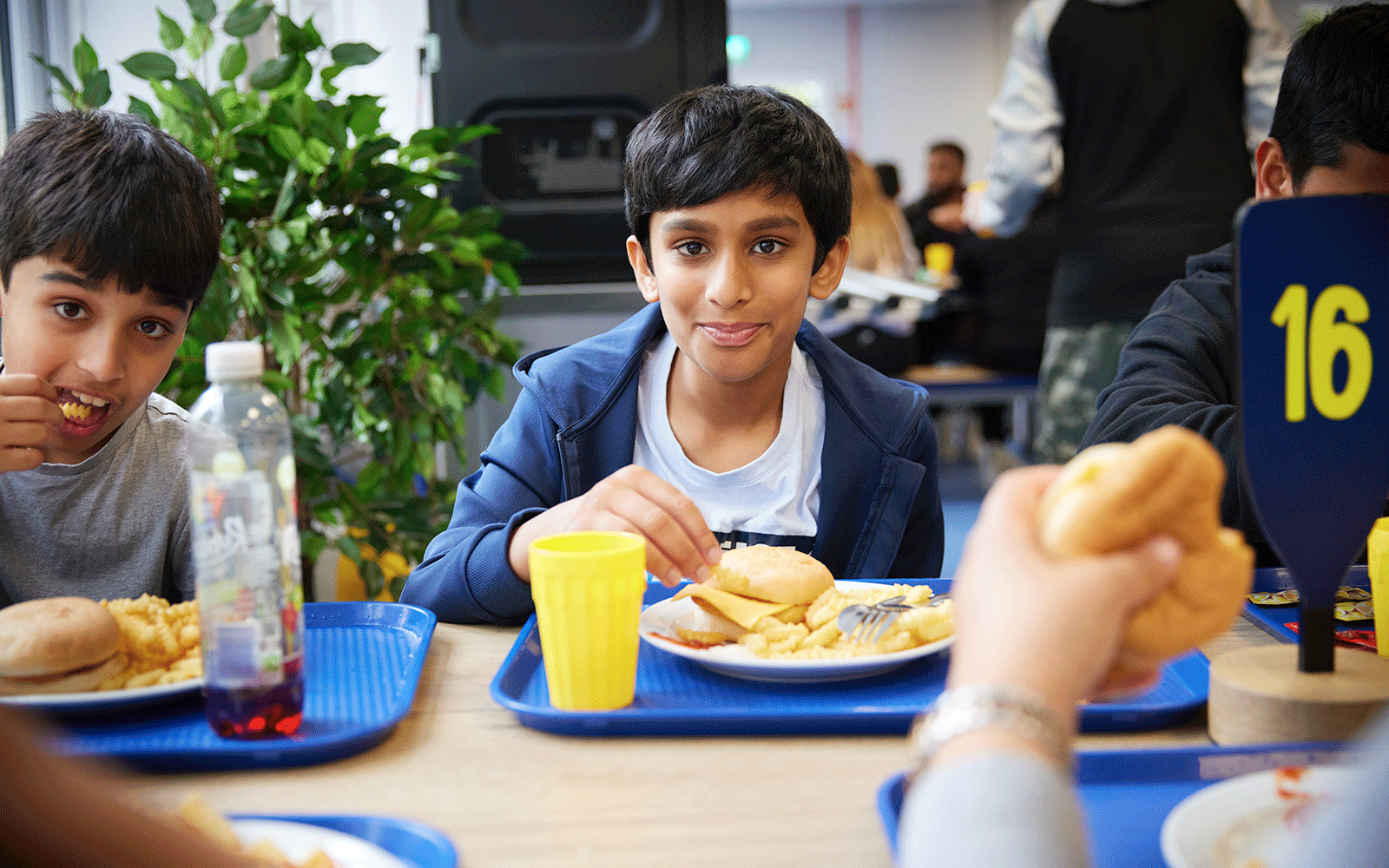 Three boys sit at a table in a cafeteria eating food from blue trays. One boy looks at the camera while holding a sandwich, and another drinks from a yellow cup. Table number "16" is visible.